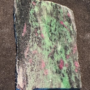 Pode incluir: Uma placa retangular de rubi em zoisite. A pedra apresenta uma mistura de cores, incluindo verde, preto e rosa. A superfície tem uma textura áspera e natural, com cristais de rubi rosa espalhados.