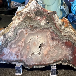 May include: A polished agate slab featuring concentric bands of translucent white, soft pink, and deep reddish-brown colors. The specimen displays a central crystalline cavity surrounded by intricate, wavy mineral layers and a rough, dark outer crust.