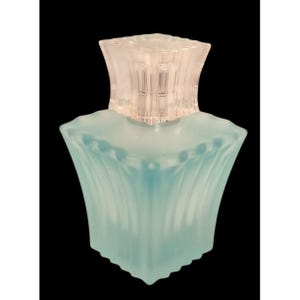 Pode incluir: Frasco de perfume de vidro azul claro com tampa transparente e canelada. O frasco tem um formato único, alargado, com linhas verticais e pequenos pontos em relevo perto do topo. O fundo é preto.