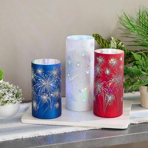 Può includere: Tre lanterne decorative cilindriche rosse, bianche e blu, ognuna ornata da un disegno di fuochi d'artificio argentati. Le lanterne sono illuminate, creando un'esposizione festosa. Sono poste su un vassoio rettangolare bianco. Misurano circa 20 cm di altezza.