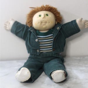 May include: A soft-sculpture doll with a round vinyl face, green eyes, and brown yarn hair. The doll wears a dark green corduroy jacket with silver snaps over a striped shirt, matching dark green corduroy pants, and white fabric shoes with brown soles.