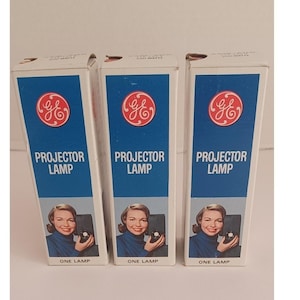 May include: Three vintage GE projector lamp boxes. Each box has a blue top with a red and white GE logo and the words "PROJECTOR LAMP". Below is a picture of a woman holding a projector. The bottom of the box reads "ONE LAMP".