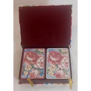 May include: A rectangular storage box with a hinged lid, featuring a deep maroon interior and a floral exterior pattern. Inside, two decks of playing cards are neatly arranged, each displaying a matching pink rose and blue floral design.