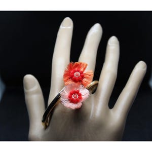 May include: A gold-tone ring featuring two flower designs. One flower is red-orange, and the other is pink. Each flower has a red center. The ring is on a mannequin hand.