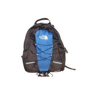 Op de afbeelding: Zwarte en blauwe rugzak met de tekst "THE NORTH FACE" in het wit. De rugzak heeft een blauw middenpaneel met kruisende bungee-koorden, een zwart handvat aan de bovenkant en zijvakken van gaas. Witte reflecterende strepen zitten aan de zijkanten.