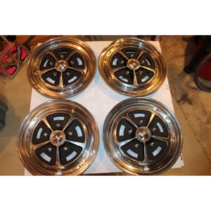 Puede incluir: Juego de cuatro llantas de coche antiguas. Cada llanta presenta una llanta plateada pulida con un centro negro y un tapacubos plateado. Las llantas están dispuestas en forma cuadrada sobre una superficie blanca.