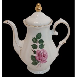 May include: White porcelain teapot with a lid and a gold-colored finial. The teapot features a pink rose and green leaves design. The handle and spout are accented with gold trim. The teapot is on a black background.
