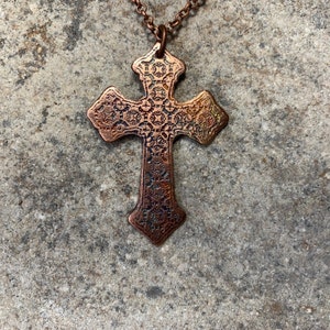 Może przedstawiać: Miedziany wisiorek w kształcie krzyża z ozdobnym, teksturowanym wzorem. Krzyż wisi na pasującym łańcuszku. Wisiorek ma szczegółowy wzór na całej powierzchni, a metal ma lekko postarzany wygląd.