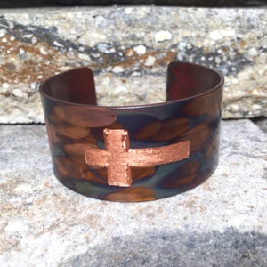 Puede incluir: Brazalete de puño de cobre con diseño de cruz. El brazalete presenta un patrón texturizado y moteado en tonos marrón, cobre y azul. La cruz es de color cobre sólido y está centrada en el brazalete.