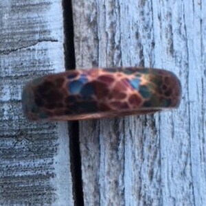 May include: A hammered copper ring with a textured surface. The ring features a mix of colors, including copper, teal, and brown. The ring is shown on a weathered wooden surface.