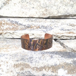 Puede incluir: Un brazalete de cobre con un patrón de corazones grabado. El brazalete tiene un aspecto rústico y envejecido con variaciones de color, incluyendo tonos de marrón, dorado y azul. El diseño abierto facilita su uso.