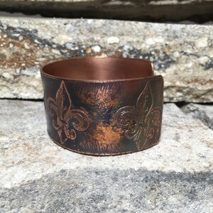 Puede incluir: Una pulsera de puño de cobre con un diseño de flor de lis. La pulsera tiene un acabado envejecido con tonos marrones y negros. El diseño de puño abierto permite un fácil uso. La pulsera mide aproximadamente 5 cm de ancho.