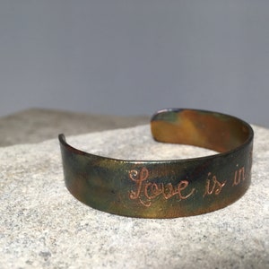 Puede incluir: Brazalete de metal hecho a mano con acabado de pátina. El brazalete está grabado con las palabras "Love is in" en fuente cursiva. El brazalete mide aproximadamente 1,9 cm de ancho.