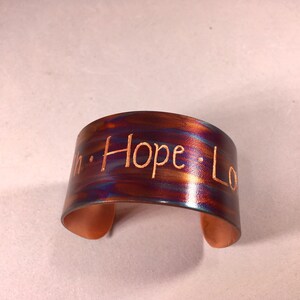 Puede incluir: Una pulsera de puño de cobre con un acabado cepillado. La pulsera tiene las palabras "Faith Hope Love" grabadas en la parte delantera. El metal tiene una mezcla de colores, incluyendo bronce, azul y morado.