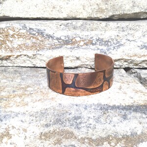 Puede incluir: Una pulsera de puño de cobre con un diseño texturizado único. La pulsera presenta un patrón oscuro e irregular sobre un fondo marrón rojizo cálido. El diseño abierto facilita su uso. La anchura de la pulsera es de aproximadamente 2,5 cm.