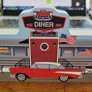 May include: A wooden model of a classic American diner with a red brick facade, a red and white car parked in front, and a sign that reads "Broad Street Diner Serving Breakfast, Lunch & Dinner".