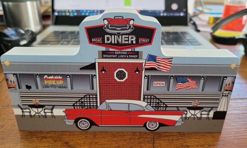 May include: A wooden model of a classic American diner with a red brick facade, a red and white car parked in front, and a sign that reads "Broad Street Diner Serving Breakfast, Lunch & Dinner".