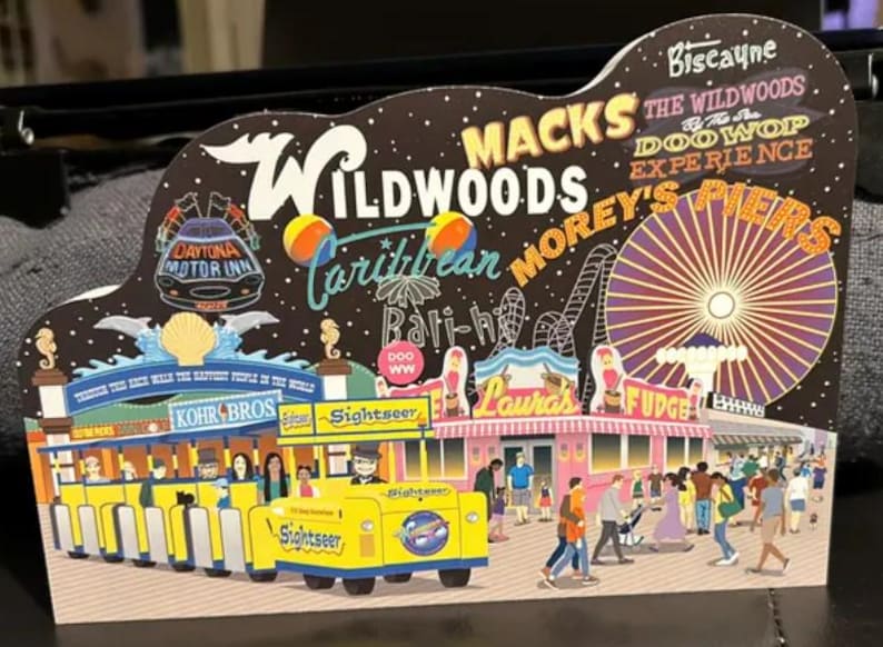 May include: A colorful illustration of a Wildwoods, New Jersey boardwalk scene with a Ferris wheel, shops, and a yellow trolley car. The text "Wildwoods" is featured in large, bold letters. The text "Mack's", "Morey's Piers", "Doo Wop Experience", and "Biscayne" are also featured in the illustration.