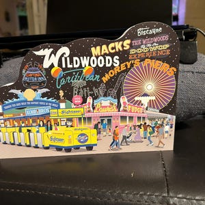 May include: A colorful illustration of a boardwalk amusement park with a Ferris wheel, shops, and a yellow and blue sightseeing train. The text "Wildwoods" is featured in the illustration.