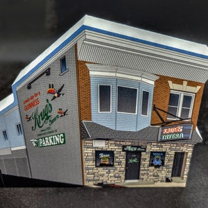 May include: A model of a brick building with a blue awning and a sign that says "Krug's Tavern". The building has a mural on the side with the text "Come in for a Guinness, Krug's Tavern" and a green sign that says "Parking".