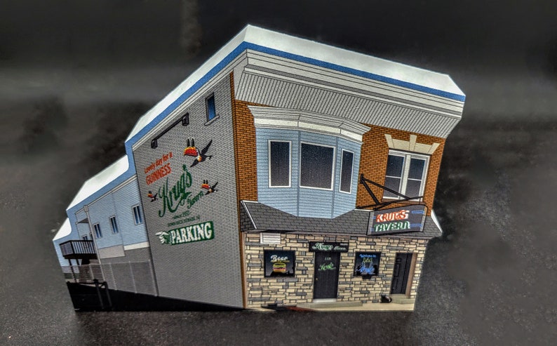 May include: A model of a brick building with a blue awning and a sign that says "Krug's Tavern". The building has a mural on the side with the text "Come in for a Guinness, Krug's Tavern" and a green sign that says "Parking".