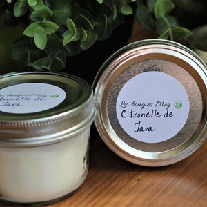 Op de afbeelding: Drie glazen potten met zilveren deksels die witte kaarsen bevatten. Elke pot heeft een wit etiket met zwarte handgeschreven tekst die "Les bougies Meg Citronelle de Java" luidt.