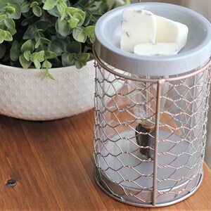 May include: A grey and silver metal wax warmer with a wire mesh cage and a white ceramic top. The warmer has a light bulb inside and is sitting on a wooden surface. There are three white wax melts on the top of the warmer.