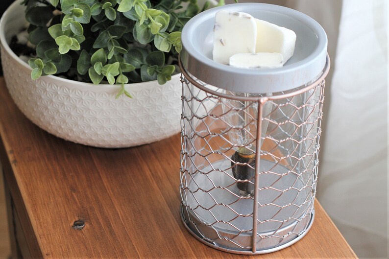 May include: A gray and silver metal wax warmer with a wire mesh cage and a white ceramic top. The warmer has a light bulb inside and is sitting on a wooden surface. There are three white wax melts on the top of the warmer.