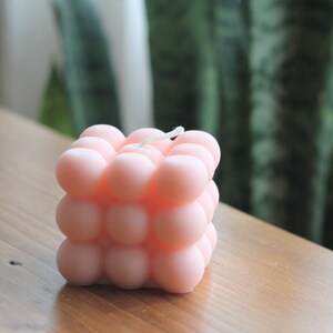 May include: A pink bubble candle with a white wick. The candle is shaped like a cube and has a textured surface.