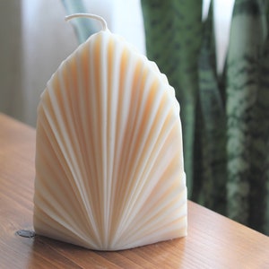 May include: A cream-coloured, fan-shaped candle with a textured, ridged design. The candle has a wick at the top and sits on a wooden surface. The candle's shape resembles a seashell.