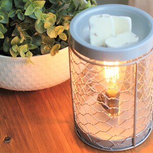 May include: A gray ceramic wax warmer with a wire cage design. The warmer has a light bulb inside and is lit. There are three white wax melts on top of the warmer.