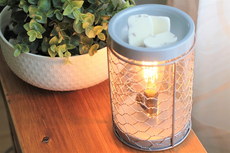 May include: A gray ceramic wax warmer with a wire cage design. The warmer has a light bulb inside and is lit. There are three white wax melts on top of the warmer.