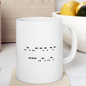 May include: White ceramic mug with a black Morse code design. The mug has a curved handle and is sitting on a beige coaster. The background includes a wooden cutting board, a white bowl with fruit, and a beige container.