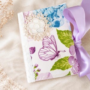May include: A fabric-covered journal featuring a white background with a purple butterfly, green leaves, and blue floral patterns. The cover is embellished with a circular cream-colored lace doily and a wide, smooth lavender satin ribbon tied on the side.