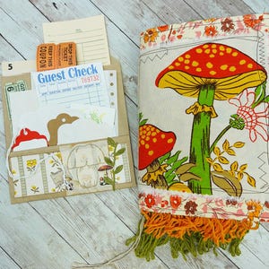 May include: A fabric-covered journal featuring a large red and yellow mushroom print with orange and green fringe at the bottom, accompanied by a tan paper folder containing various paper ephemera, including a guest check and orange tickets.