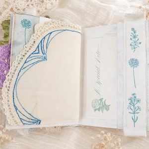 May include: An open journal featuring a cream-colored page with a blue embroidered lace corner accent. The opposite page displays a white card with the text A Special Note and a rose illustration, alongside a fabric strip with blue floral prints.