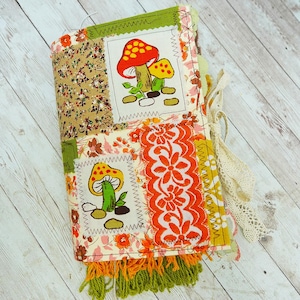 May include: A fabric-covered journal featuring a patchwork design with floral patterns, lace trim, and two rectangular patches depicting colorful mushrooms. The spine and bottom edge are decorated with green and orange fringe and cream lace.