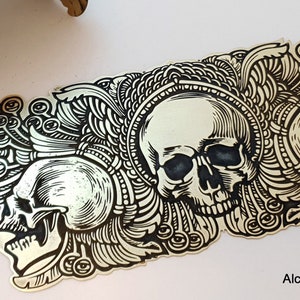 May include: A gold metal cuff bracelet featuring a black and white skull design. The bracelet is adorned with intricate details and has a curved shape. The text "Alchimia Metal Magic - Art Studio" is visible at the bottom of the image.
