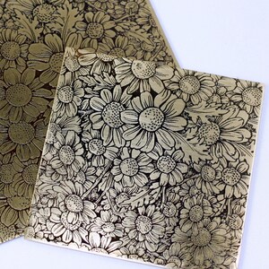 May include: Two gold metal sheets with a raised embossed design of black daisies. The sheets are different sizes and are lying on a white surface.