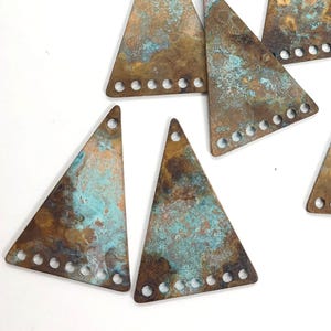 May include: A collection of ten triangular metal pendants with a distressed, aged appearance. The pendants feature a mix of brown, copper, and turquoise colors, with small holes along the bottom edge and a single hole at the top for stringing.