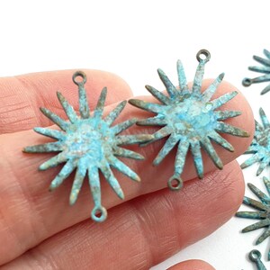 May include: A close-up of a hand holding a group of turquoise blue metal starburst charms with a loop at the top. The charms are all the same size and have a rustic, aged look.