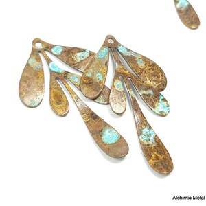 May include: Close-up of several metal teardrop-shaped pendants with a distressed, aged appearance. The pendants are primarily bronze with areas of turquoise-colored patina. Each has a small hole at the top for stringing.
