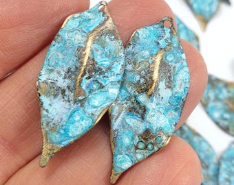 May include: Close-up of several leaf-shaped metal charms. The charms have a turquoise and bronze patina, with a small hole at the top for stringing. The leaves are approximately 1.5 inches long.