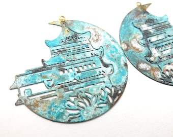 May include: Two metal, circular, decorative ornaments with a cut-out design of a pagoda and foliage. The ornaments are painted in shades of blue, green, brown, and white, giving them a distressed, aged look.