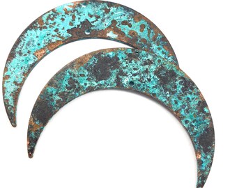 May include: Two crescent moon-shaped metal components featuring a weathered, oxidized finish with mottled patches of turquoise, teal, and dark brown rust. Each piece has two small circular holes near the top edge for attachment purposes.