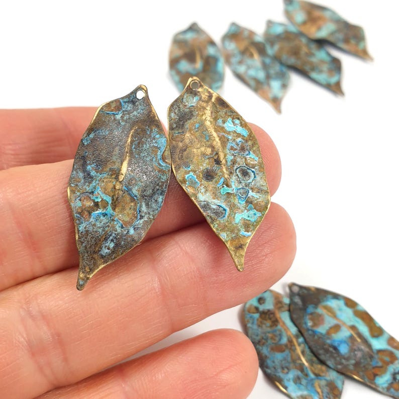 May include: A collection of leaf-shaped metal charms featuring a weathered, oxidized finish with mottled patches of metallic bronze, dark brown, and bright turquoise blue. Each charm has a small circular hole at the top for attachment purposes.