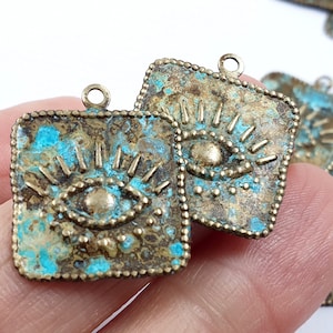 Może przedstawiać: Dwa kwadratowe charms z mosiądzu z wzorem oka. Charms mają postarzaną, zużytą powierzchnię z turkusową patyną. Każdy charm ma małą pętelkę u góry do przymocowania do naszyjnika lub bransoletki.