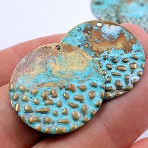 May include: Two round, textured metal charms with a turquoise and gold patina. The charms have a hole at the top for hanging.
