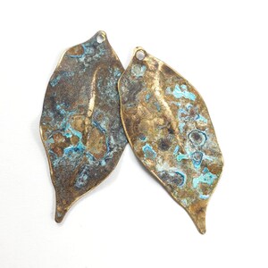 May include: Two leaf-shaped metal pendants featuring a textured, weathered surface with a mix of metallic bronze and oxidized turquoise blue patina. Each piece has an irregular, organic edge and a small circular loop at the top for attachment.