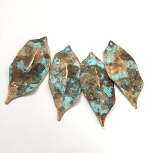May include: Four leaf-shaped metal charms featuring a weathered, oxidized finish with a mottled blend of metallic bronze, brown, and vibrant turquoise blue patina. Each piece has a small circular hole at the top for attachment and a textured surface.